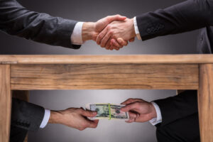 Close-up Of Two Businesspeople Shaking Hand And Taking Bribe Under Wooden Table On Grey Background, representing TRICARE bribery