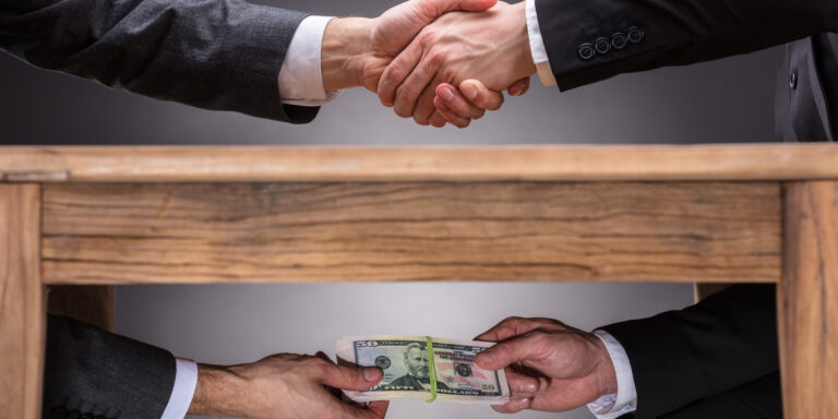Close-up Of Two Businesspeople Shaking Hand And Taking Bribe Under Wooden Table On Grey Background, representing TRICARE bribery